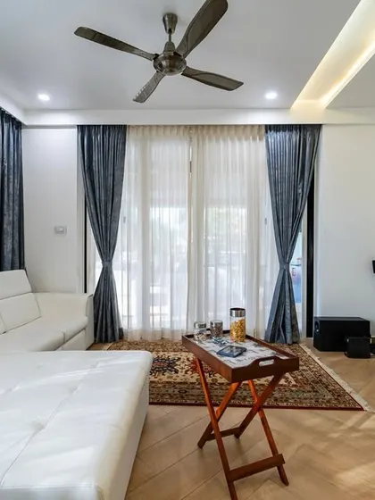 This spacious bedroom combines a living area with a sleeping space. A chic white sectional sofa faces a TV set against a striking geometric accent wall, creating a cozy retreat for relaxing or watching movies.