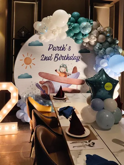 An airplane-themed 2nd birthday for little Parth! This setup included a custom-printed backdrop with a cute pilot illustration, a balloon arch in sky-blue and white, and a marquee number '2'.
