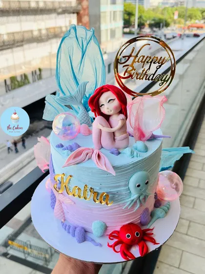 An enchanting Little Mermaid cake featuring a hand-sculpted Ariel. The design uses a blend of blue and pink buttercream, with fondant sea creatures like an octopus and crab to complete the underwater scene.
