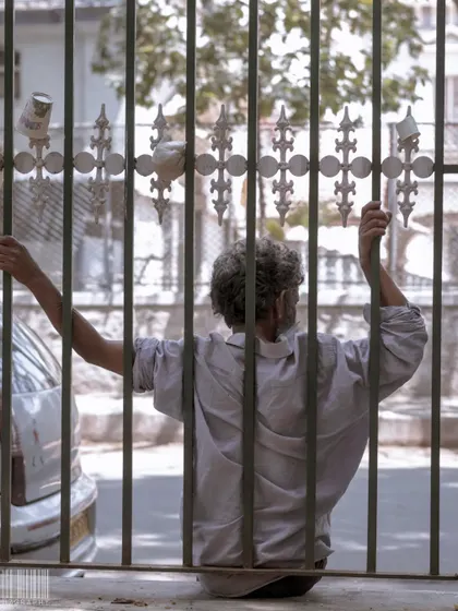 A man looking out from behind a gate. The composition uses the bars as a frame, creating a feeling of being confined or waiting.
