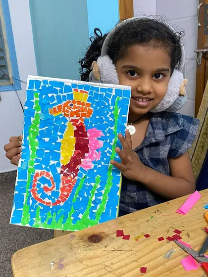 A proud artist shows off her seahorse mosaic. Notice her noise-canceling earmuffs, an example of how we adapt our classroom to be a sensory-friendly space for every child's needs.