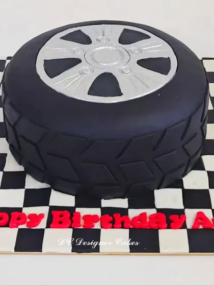 This car tyre cake was designed for a car lover's birthday. The realistic tread and silver rim are crafted from fondant, sitting on a checkered flag board to complete the racing theme.