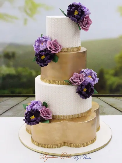 A grand four-tier wedding cake combining white stenciled tiers with brushed gold tiers. The design is brought together by cascading clusters of handmade sugar flowers in shades of lilac and purple.