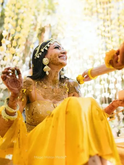 The bride glows with joy during her Haldi ceremony, covered in turmeric paste. The background of hanging florals adds to the festive and beautiful moment.