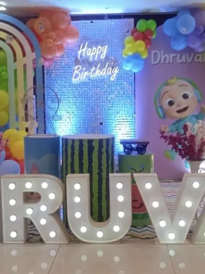 A Cocomelon themed first birthday with large illuminated letters, character cutouts, and a backdrop featuring a blue shimmer panel.
