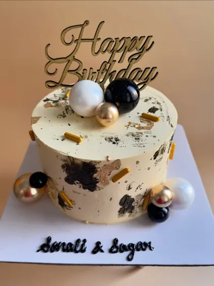 A sophisticated birthday cake with a textured black and gold foil design on a cream-colored base. The look is completed with a clean arrangement of black, white, and gold spheres.
