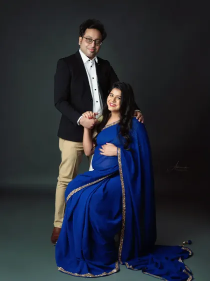 A formal and elegant couple's portrait. The mom-to-be is seated, draped in a beautiful blue saree, while her partner stands by her side.