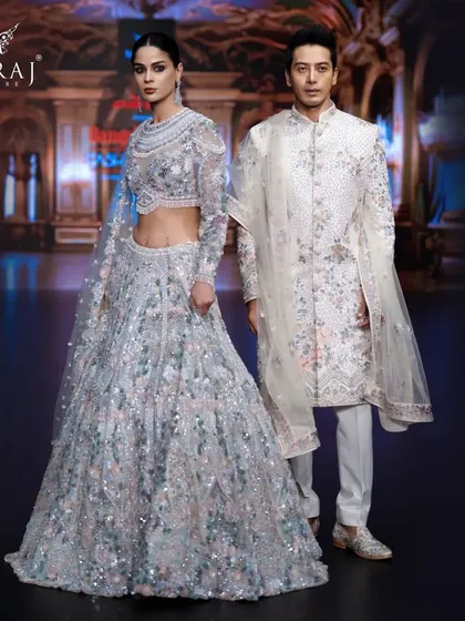 A stunning bridal couple graces the stage in an off-white bridal lehenga and men's sherwani. The groom's attire is adorned with exquisite floral multicolor embellishments, showcasing timeless elegance.