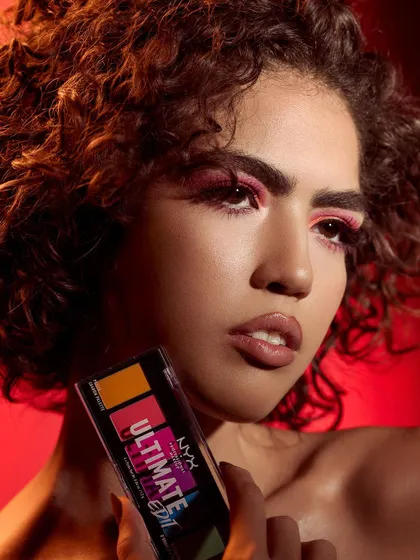 A more intense and focused shot from the NYX campaign. The model holds the palette, creating a direct link to the bold pink eyeshadow look. The red background adds to the high-energy, dramatic feel.