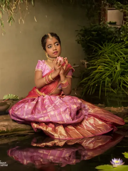A girl in a traditional silk lehenga sits by a pond, holding a lotus flower. The reflection in the water adds a beautiful, serene touch.