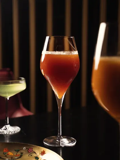 An elegant, tall glass of a reddish-brown beverage, shot in a dimly lit, fine-dining setting. The focus is on the glassware and the deep color of the drink, conveying sophistication.