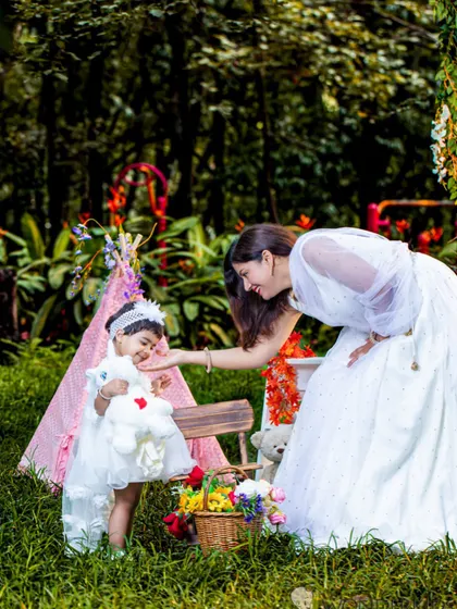 A mother and daughter share a tender moment during an outdoor photoshoot. The beautiful white dresses and the lush green background create a serene and angelic scene.