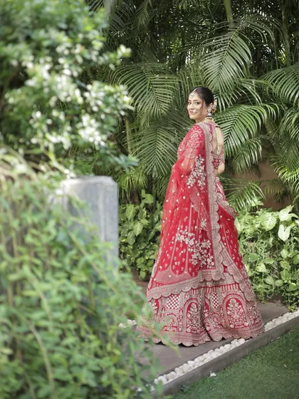 A full-length shot of the bride in her magnificent red lehenga, showcasing the complete look in a beautiful garden setting.