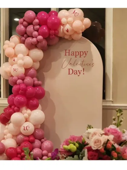A beautiful Valentine's Day setup featuring a white arched panel and a balloon garland in various shades of pink and rose gold. The custom lettering and lipstick kiss marks add a personal touch.
