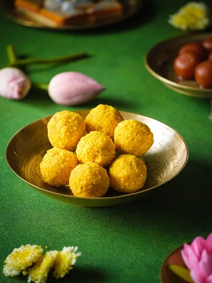 A close-up of a bowl of ladoos on a green background, surrounded by lotus flowers, creating a vibrant and traditional composition.