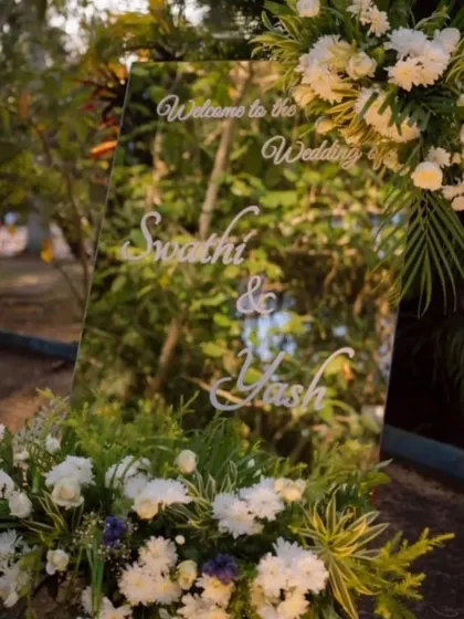 A modern and elegant welcome sign on a mirror, framed with white flowers, reflecting the natural beauty of the outdoor venue.