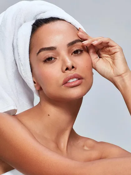 A classic "fresh out of the shower" beauty portrait for a skincare brand. The model, with her hair in a white towel, has a clean, natural look that emphasizes healthy, well-cared-for skin.