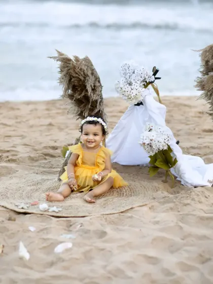 A sweet photo of a baby girl sitting on a jute mat on the beach for her first birthday, with the waves gently rolling in behind her.