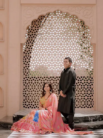 A classic portrait of the couple against an intricate jaali wall, showcasing their traditional outfits and the grandeur of the location.
