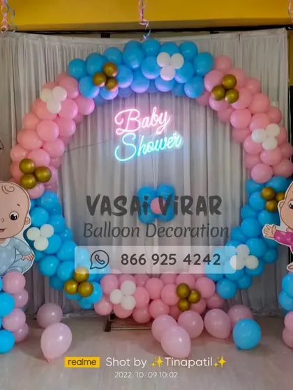 This is a full view of the popular ring backdrop for a baby shower. The circular frame is covered in pink and blue balloons with gold accents, and flanked by cute baby cutouts and "BABY" letter blocks.
