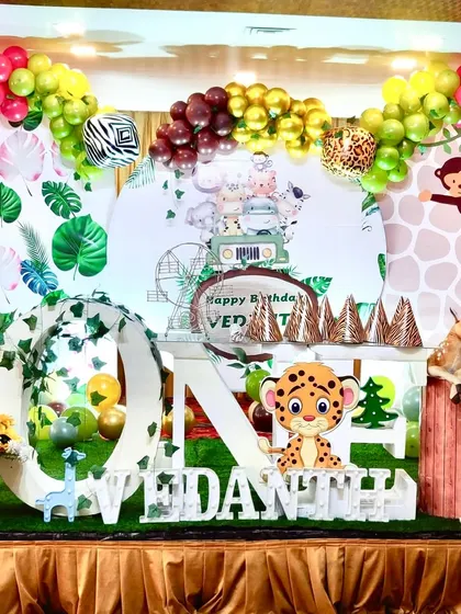 A complete stage view of a jungle-themed first birthday party. This setup features a large "ONE" display, a personalized backdrop, and an elaborate balloon garland in shades of green, yellow, and brown with animal print details.