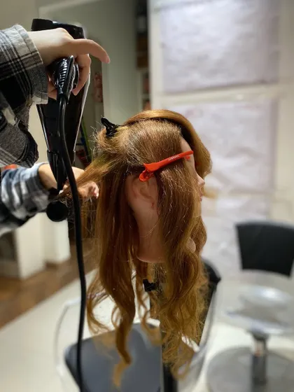 A student practices her blow-drying technique, focusing on creating lift at the root and smoothness through the ends.