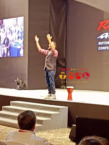 The facilitator stands on stage, arms raised, inviting the audience to join in. This gesture of openness is key to our philosophy of making everyone feel welcome and part of the experience.