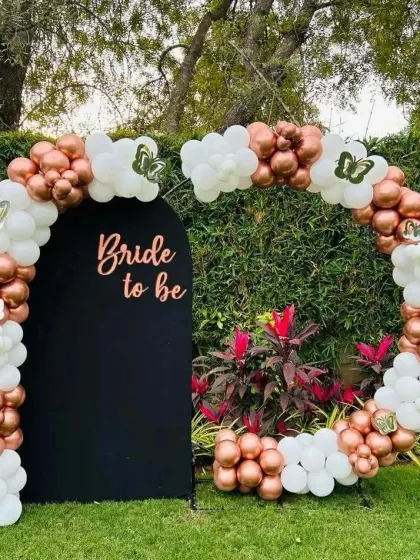 An elegant outdoor "Bride to be" setup with a black arch backdrop and a balloon garland of white and rose gold.