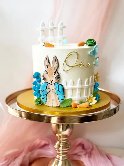 A charming Peter Rabbit cake for a first birthday. This design features a hand-painted Peter Rabbit, a white picket fence, and delicate fondant flowers and carrots.
