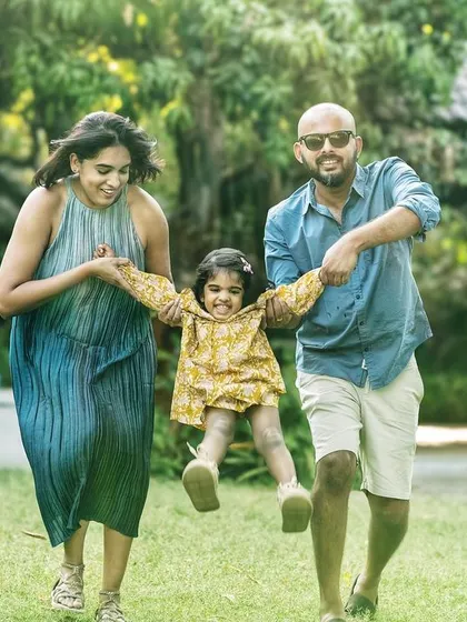 Pure, joyful motion. A mother and father swing their daughter between them as they run through a grassy park, a perfect picture of carefree family fun.