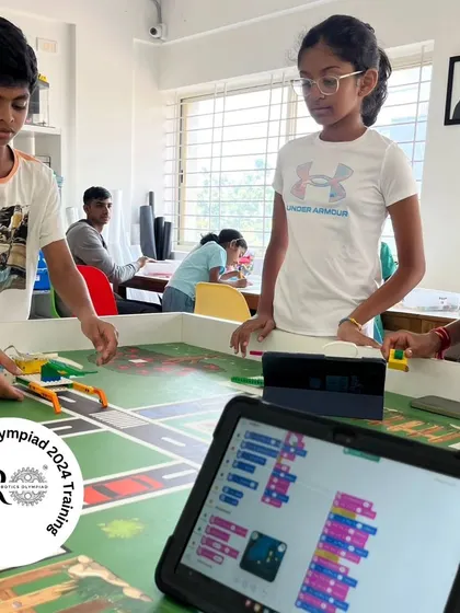 A team of students collaborates on their strategy for the IRO 2024. The tablet in the foreground displays the block-based code they are writing to control their robot's actions on the farm-themed mat.