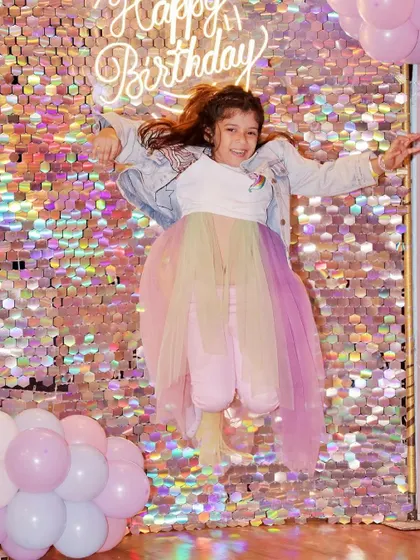 The birthday girl jumps for joy in front of a shimmering, iridescent sequin wall at her unicorn-themed party.