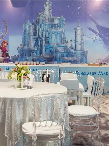 The dining area for the 'Frozen' party, with tables and chairs draped in white to resemble a snowy landscape. The backdrop features a panoramic view of Elsa's ice castle, fully immersing the children in the theme.