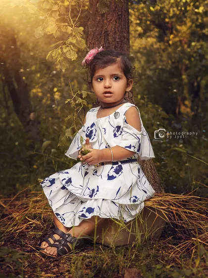 A sweet portrait of a toddler girl sitting at the base of a tree in a sunlit forest. The natural setting provides a beautiful and peaceful backdrop.