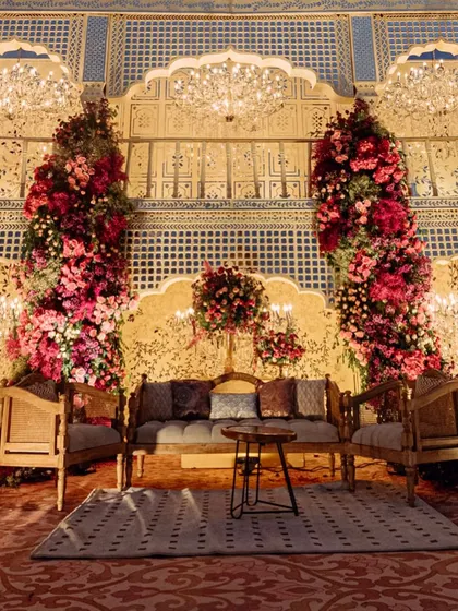 A seating area for guests, featuring a backdrop of intricate jali screens and towering floral arrangements. The design provides both comfort and a visually stunning environment.