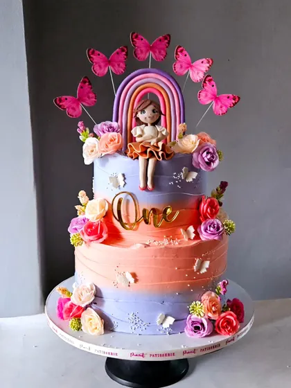 A glorious first birthday cake for a little princess. This three-tiered cake features a beautiful peach and lavender ombre effect, adorned with fresh flowers, butterflies, and a custom girl figurine.