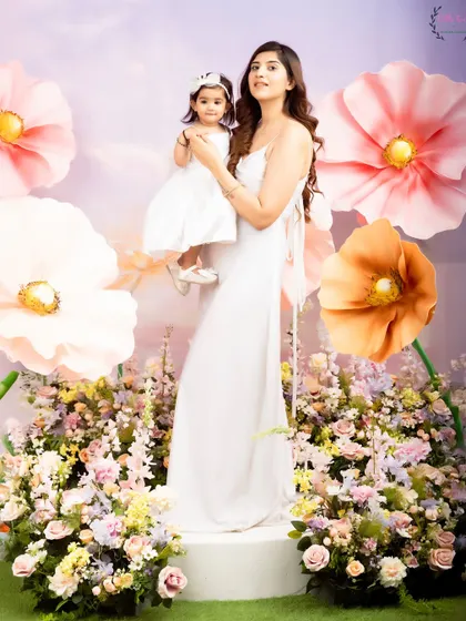 A full-length portrait of a mother and daughter in a beautiful, flower-filled studio setup. Their matching white dresses and the whimsical background create a stunning, high-fashion-inspired image.