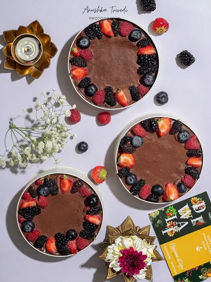 A flat lay of three round chocolate cakes topped with fresh berries, styled with Diwali diyas for a festive fusion.