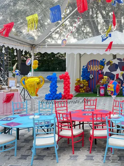 The seating area for a soccer-themed 11th birthday party. The tables and chairs are in a vibrant red and blue, matching the Barcelona team colors, and the space is decorated with a trophy balloon and team banners.