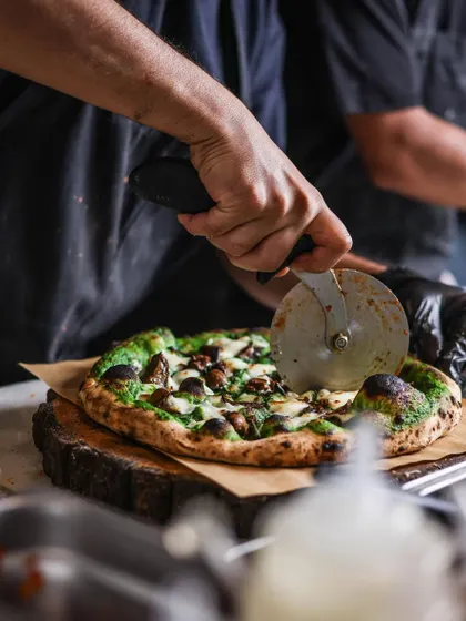 Slicing up a green Fungi pizza. This action shot gives a great view of the toppings, including mushrooms and creamy burrata, on a spinach pesto base.
