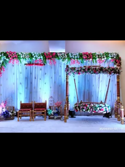 A classic naming ceremony or engagement setup with a beautifully decorated floral swing (jhula). The stage includes seating for guests and is set against a simple white curtain backdrop with a floral border.