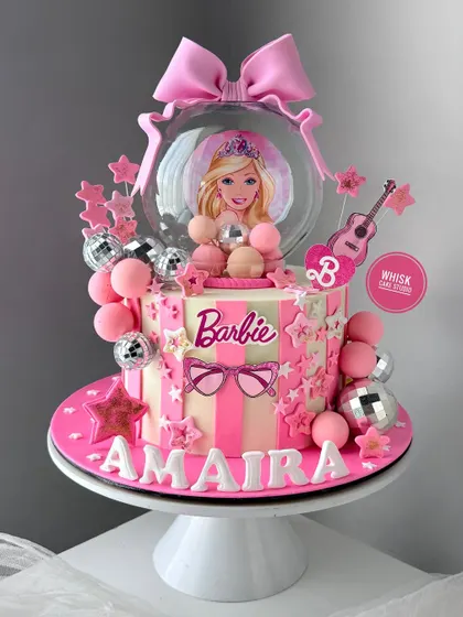 A fun and glamorous Barbie-themed cake. This design features a Barbie snow globe, disco balls, and lots of pink stars for a true Barbie girl.