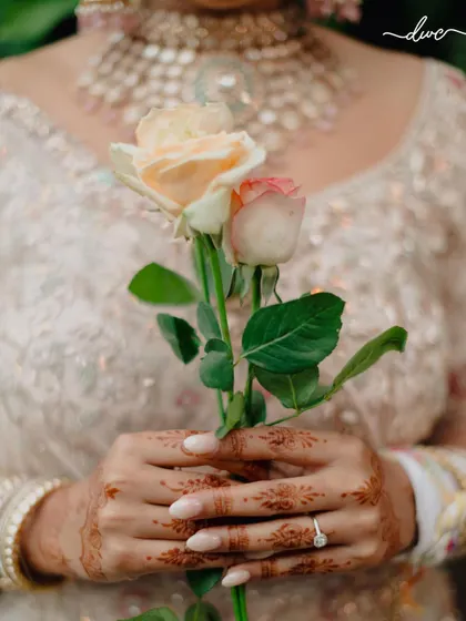 A close-up detail shot of the bride's hands, adorned with mehendi and a delicate engagement ring, holding two beautiful roses.