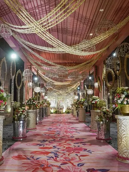 A beautiful floral-printed carpet creates a vibrant aisle, flanked by elegant pedestals with flower arrangements. The ceiling is adorned with draped fabric and traditional pearl garlands, creating a rich, layered look.