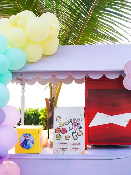 A pastel purple popcorn cart set up at a beachside Disney Princess party, decorated with a soft rainbow balloon garland.