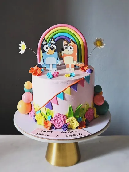 A colorful and happy Bluey-themed cake for a joint birthday. This design features Bluey and Bingo under a pastel rainbow, with festive bunting and a fun, textured floral border.