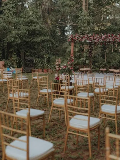 An outdoor ceremony setup in a beautiful garden, with golden chairs and a rustic floral mandap, creating a warm and inviting atmosphere.