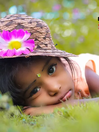 A close-up portrait of a young girl lying in the grass. Her curious eyes and the pretty flower on her hat are the focus, creating a gentle and innocent image.
