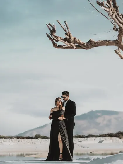 A dramatic and modern pre-wedding portrait set against a stark, beautiful landscape. The composition uses a bare tree to frame the couple, creating a powerful, high-fashion image that feels both artistic and romantic.