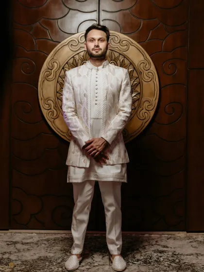 A dapper portrait of the groom in his elegant white sherwani, posing against a beautifully carved wooden backdrop before his roka ceremony.